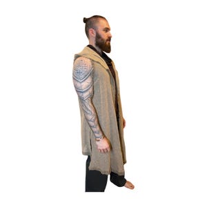 May include: A long, hooded, open-front cardigan in a neutral tan color. The cardigan has a relaxed fit and a split hem. The person in the image is wearing black pants and a dark shirt underneath the cardigan.