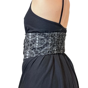 May include: Black dress with spaghetti straps and a wide, patterned belt. The belt features a repeating white design on a black background. The dress has a fitted bodice and a flowing skirt, creating a flattering silhouette.