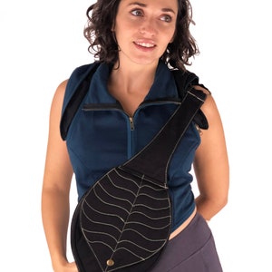 May include: A black leaf-shaped crossbody bag with a gold button. The bag is worn by a person wearing a blue sleeveless shirt and gray pants.