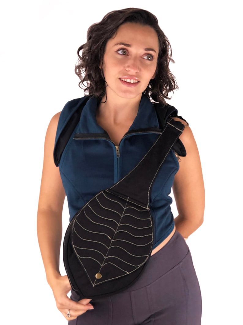 May include: A black leaf-shaped crossbody bag with a gold button. The bag is worn by a person wearing a blue sleeveless shirt and gray pants.