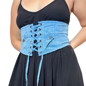 May include: Light blue denim corset with a lace-up front and button details. The corset is worn over a black dress. The corset has a faded wash and a structured design, offering a unique style.