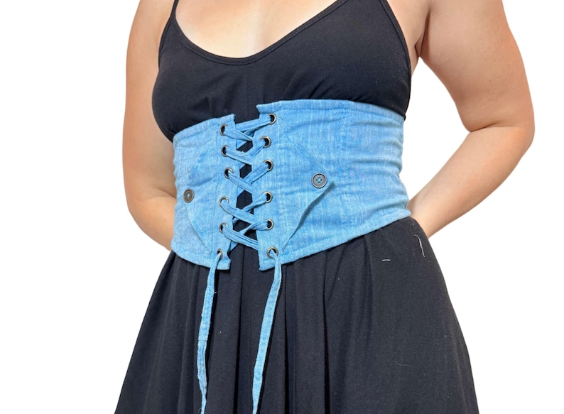 May include: Light blue denim corset with a lace-up front and button details. The corset is worn over a black dress. The corset has a faded wash and a structured design, offering a unique style.