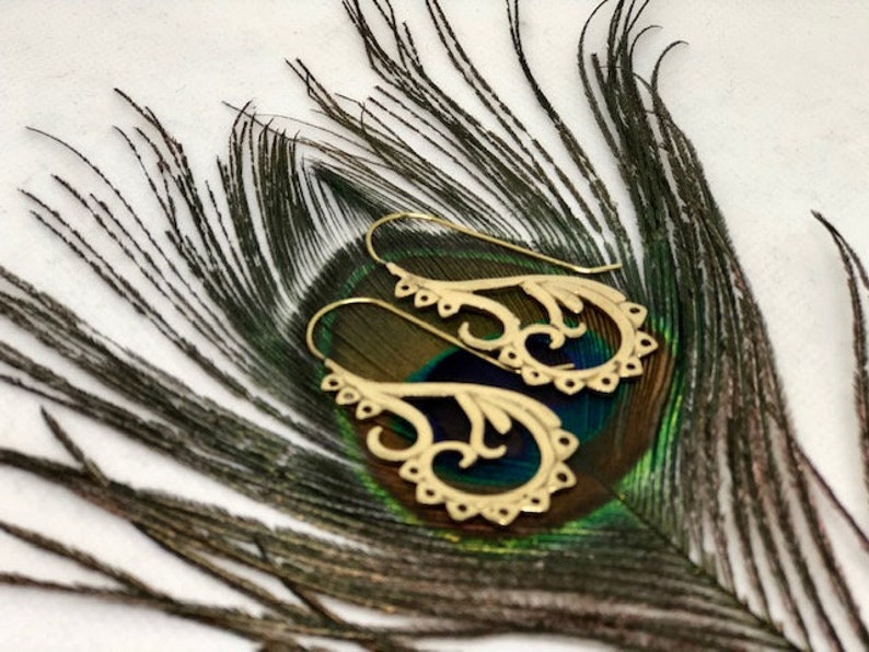 May include: A pair of gold-toned earrings with a delicate, intricate design. The earrings are shaped like a stylized flower with a curved stem. They are displayed on a peacock feather.