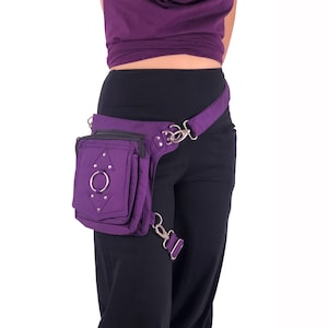 May include: Purple fanny pack with a silver ring and studs. The pack has a zippered pocket and a strap that goes around the waist and over the shoulder.