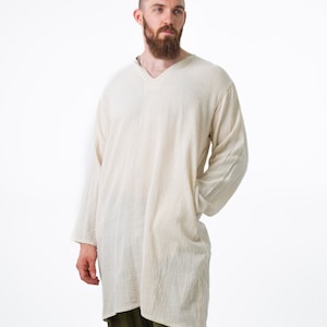 May include: A man wearing a long-sleeved, off-white tunic with a v-neck and a pair of olive green pants.