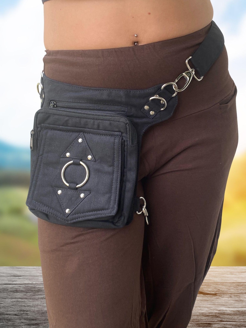 May include: A black canvas fanny pack with a silver ring and a triangle design. The fanny pack is worn on the hip and has a strap that goes around the waist.