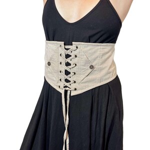 May include: Beige corset belt with a lace-up front and two button details. The corset is made of a textured fabric and is worn over a black dress. The belt cinches at the waist.