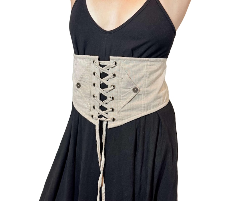 May include: Beige corset belt with a lace-up front and two button details. The corset is made of a textured fabric and is worn over a black dress. The belt cinches at the waist.