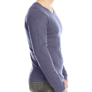 May include: A long-sleeved, V-neck shirt in a muted blue hue. The shirt features a geometric stitched design on the shoulders and down the sides. The model is wearing blue jeans.