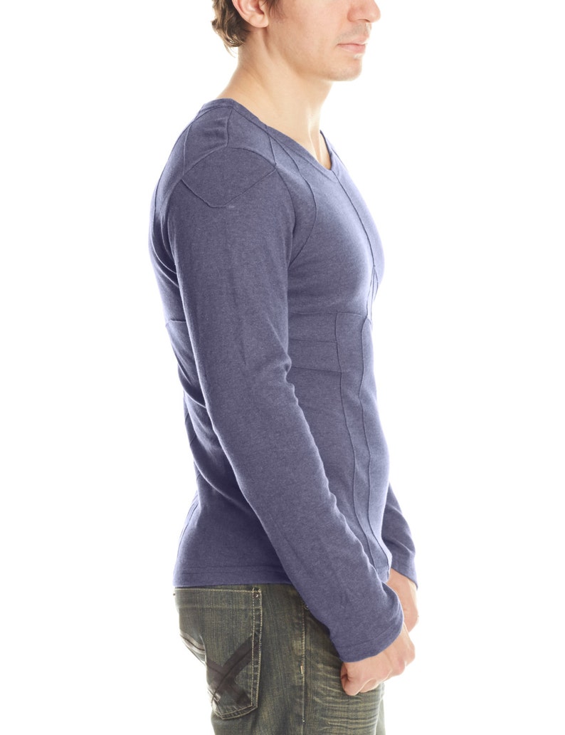 May include: A long-sleeved, V-neck shirt in a muted blue hue. The shirt features a geometric stitched design on the shoulders and down the sides. The model is wearing blue jeans.
