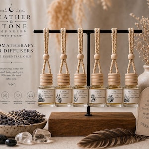 May include: Aromatherapy car diffusers with essential oils. The image shows six small glass bottles hanging from a black stand. Each bottle has a wooden top and a braided cord. The bottles are labeled with different scents and botanical illustrations. A bowl of lavender, crystals, and a feather are also visible.