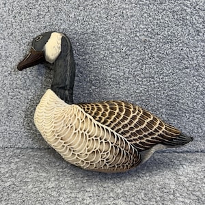 May include: A detailed carving of a Canada goose. The goose features a black head and neck, a white cheek patch, and a brown beak. The body is a mix of brown and cream-colored feathers, with intricate detailing. The goose is a decorative item.
