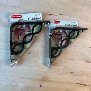 Könnte beinhalten: Zwei 15 cm Celtic Scroll-Winkel von Rubbermaid. Die Winkel sind dunkelbraun mit einem dekorativen keltischen Knotenmuster. Jeder Winkel ist auf einer Kartonrückseite mit Produktname und Marke verpackt.