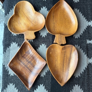 Pode incluir: Quatro travessas de madeira em forma de naipes de cartas: um trevo, uma espadilha, um diamante e um coração. As travessas são feitas de madeira castanha clara e têm um acabamento liso. As travessas estão dispostas sobre uma superfície estampada.