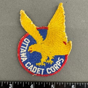 May include: A circular embroidered patch featuring a yellow eagle in flight against a blue background, bordered by a red edge. The text OTTAWA CADET CORPS is stitched in white along the bottom curve. A ruler shows the patch is 3 inches wide.