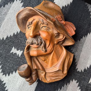 May include: A carved wooden wall hanging depicting an elderly man with a mustache and a hat, holding a pipe. The carving is in shades of brown and orange, with intricate details. The man's face shows wrinkles and a thoughtful expression. The background is a black and white patterned fabric.