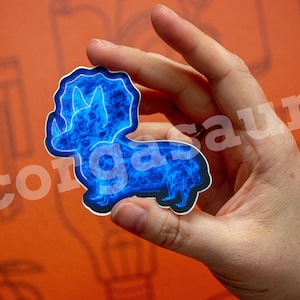May include: A blue and black sticker of a corgi dog with a triceratops head and a blue flame design. The sticker says "corgasaur" on it.