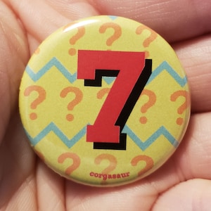 May include: A yellow button with a red number 7 on it. The button has a pattern of blue and yellow zigzags and orange question marks. The text "corgasaur" is printed on the bottom of the button.