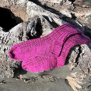 May include: A pair of vibrant fuchsia-colored knitted socks. The socks are made of a textured yarn with a ribbed pattern on the top and a patterned design on the foot. They are resting on a weathered, gray driftwood log.
