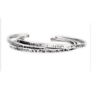 May include: A set of three silver-colored metal cuff bracelets featuring a textured, hammered finish. Each bracelet has an open-ended design with a slightly irregular, organic surface pattern that catches the light across the metallic bands.