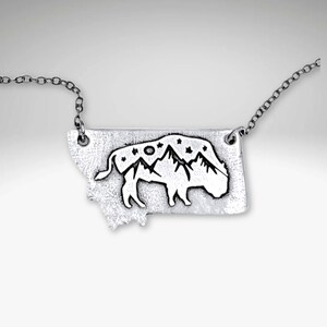 May include: Silver necklace featuring a pendant shaped like the state of Montana with a silhouette of a bison and mountains inside. The pendant is decorated with stars and a moon.