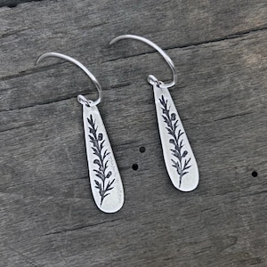 May include: A pair of silver-toned dangle earrings. Each earring features a teardrop-shaped pendant with a black botanical design. The earrings hang from a curved wire. The earrings are displayed on a weathered wooden surface.