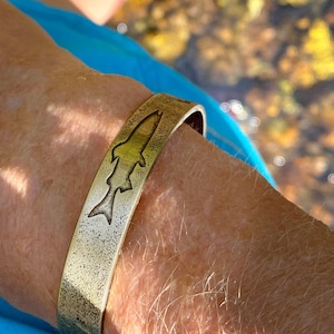 May include: A gold-colored metal cuff bracelet with a fish silhouette design. The bracelet has a textured surface and is worn on a wrist. The fish design is etched into the metal.