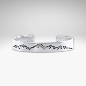 May include: A silver cuff bracelet with a mountain range design. The bracelet is made of a brushed metal and features a dark gray mountain silhouette across the front. The open cuff design allows for easy wear.