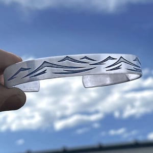 May include: A silver bracelet with a mountain range design etched into the surface. The bracelet is open and has a smooth, polished finish.