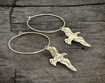 May include: A pair of gold-toned hoop earrings, each featuring a dangling charm shaped like a bird in flight. The bird charms have a textured, hammered surface finish. The thin, circular hoops are simple and open, providing a lightweight appearance.