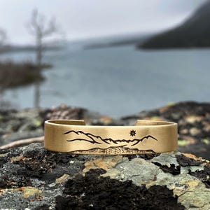 May include: A gold-colored cuff bracelet with a mountain range and sun design. The open cuff bracelet is resting on a dark, textured rock. The background features a body of water and a cloudy sky.