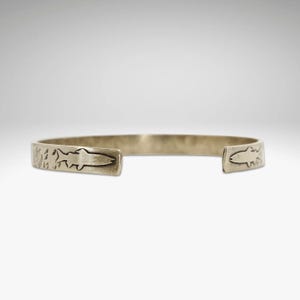 May include: A gold-toned metal bracelet with a fish design on each end. The bracelet is textured and has a hammered finish.