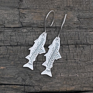 May include: A pair of silver-toned fish-shaped dangle earrings featuring etched details, including a lateral line and a pattern of small dots along the body, suspended from curved metal ear wires for a secure fit and a distinct appearance.
