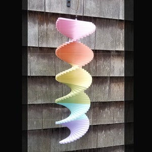 May include: A rainbow-colored, multi-layered, spiral wind spinner hanging from a hook on a wooden wall. The wind spinner is made of thin, overlapping pieces of wood.