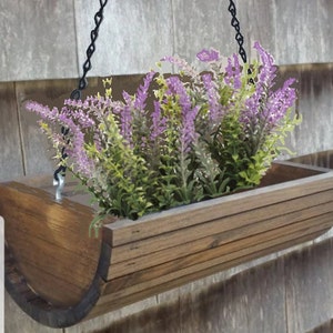 May include: A wooden hanging planter with a curved shape and a chain for hanging. The planter is filled with purple and green artificial flowers.
