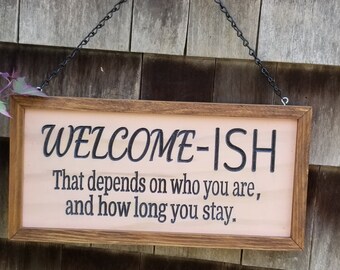 May include: A wooden sign with the text "WELCOME-ISH That depends on who you are, and how long you stay." hanging on a wooden wall.