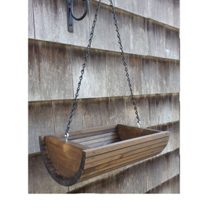 May include: A wooden hanging planter with a rounded shape and a dark brown stain. The planter is suspended from a black chain attached to a black metal hook.