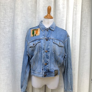 May include: Light blue denim jacket with a patch that says "IRELAND" in green, white, and orange. The jacket has two chest pockets, button closures, and a small patch on the lower right side. The jacket is displayed on a mannequin.