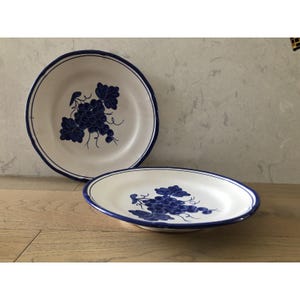 May include: Two white ceramic plates with a blue grape design. One plate is round, and the other is oval. Both plates have a blue border and a hand-painted grape cluster in the center. The plates are on a wooden surface.