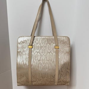 May include: A beige tote bag with a snakeskin pattern. The bag has two long straps and gold-colored accents. The bag is rectangular and has a structured design, suitable for carrying various items.