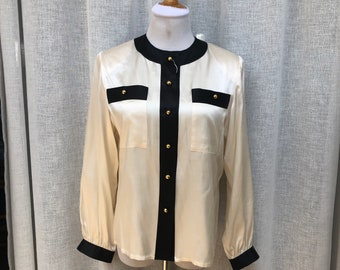 May include: Cream-colored long-sleeve blouse with black trim and gold buttons. The blouse features a round neckline, two front pockets, and black cuffs. The garment is displayed on a mannequin against a neutral backdrop.