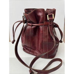May include: A burgundy leather bucket bag with a drawstring closure and a long, adjustable shoulder strap. The bag features vertical stitching details and a gold-colored buckle. The leather has a slightly distressed appearance.
