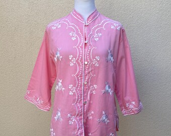 May include: A pink button-up shirt with white embroidery of flowers and leaves. The shirt has a mandarin collar and three-quarter sleeves.
