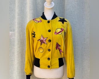 May include: A yellow velvet bomber jacket with black trim and black buttons. The jacket is decorated with sequined patches of stars, planets, and arrows. The jacket is displayed on a mannequin.