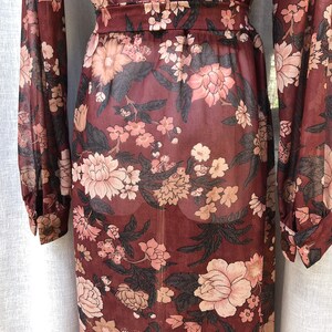 K&ouml;nnte beinhalten: Ein lang&auml;rmeliges, durchsichtiges Maxikleid mit einem Blumendruck in Braun-, Rosa- und Schwarzt&ouml;nen. Das Kleid hat einen figurbetonten Oberteil und einen flie&szlig;enden Rock.