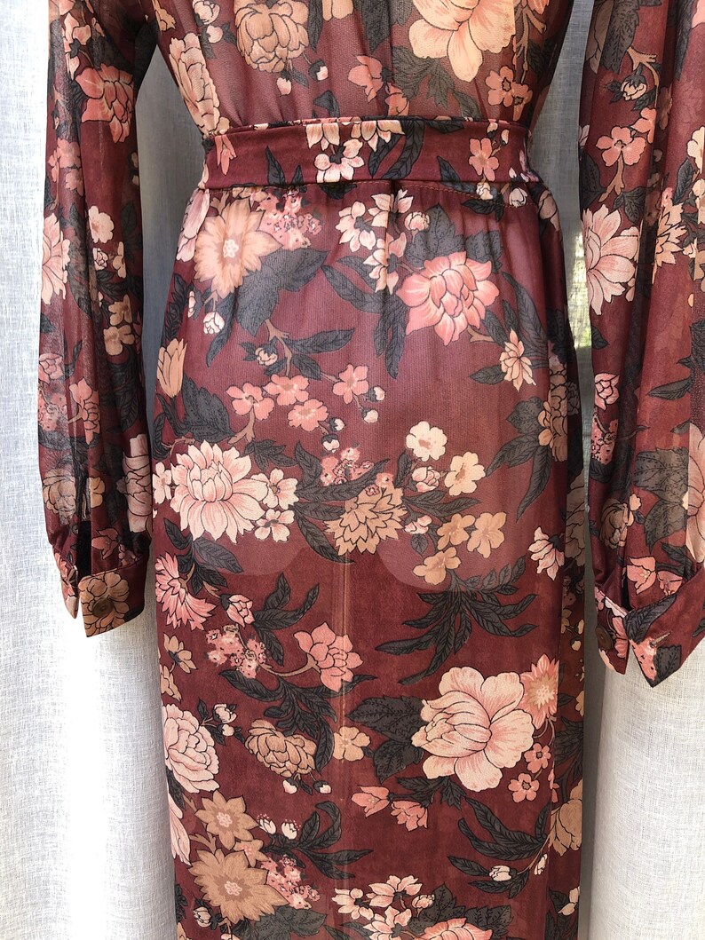 K&ouml;nnte beinhalten: Ein lang&auml;rmeliges, durchsichtiges Maxikleid mit einem Blumendruck in Braun-, Rosa- und Schwarzt&ouml;nen. Das Kleid hat einen figurbetonten Oberteil und einen flie&szlig;enden Rock.