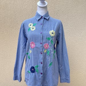 May include: A blue and white gingham button-up shirt with embroidered flowers in shades of pink, yellow, and green.