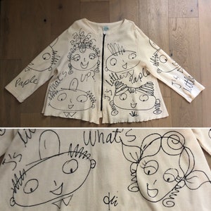 May include: A cream-colored cardigan with a zipper closure and black hand-drawn illustrations of faces and text. The text includes "What's Up?", "It's Little", "People", and "J's Little".