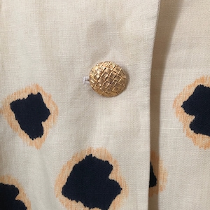 May include: A close-up of a beige linen blazer with a woven gold button. The blazer has a black and orange abstract print.