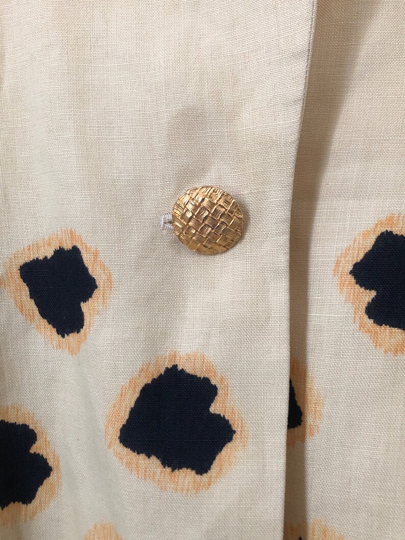 May include: A close-up of a beige linen blazer with a woven gold button. The blazer has a black and orange abstract print.